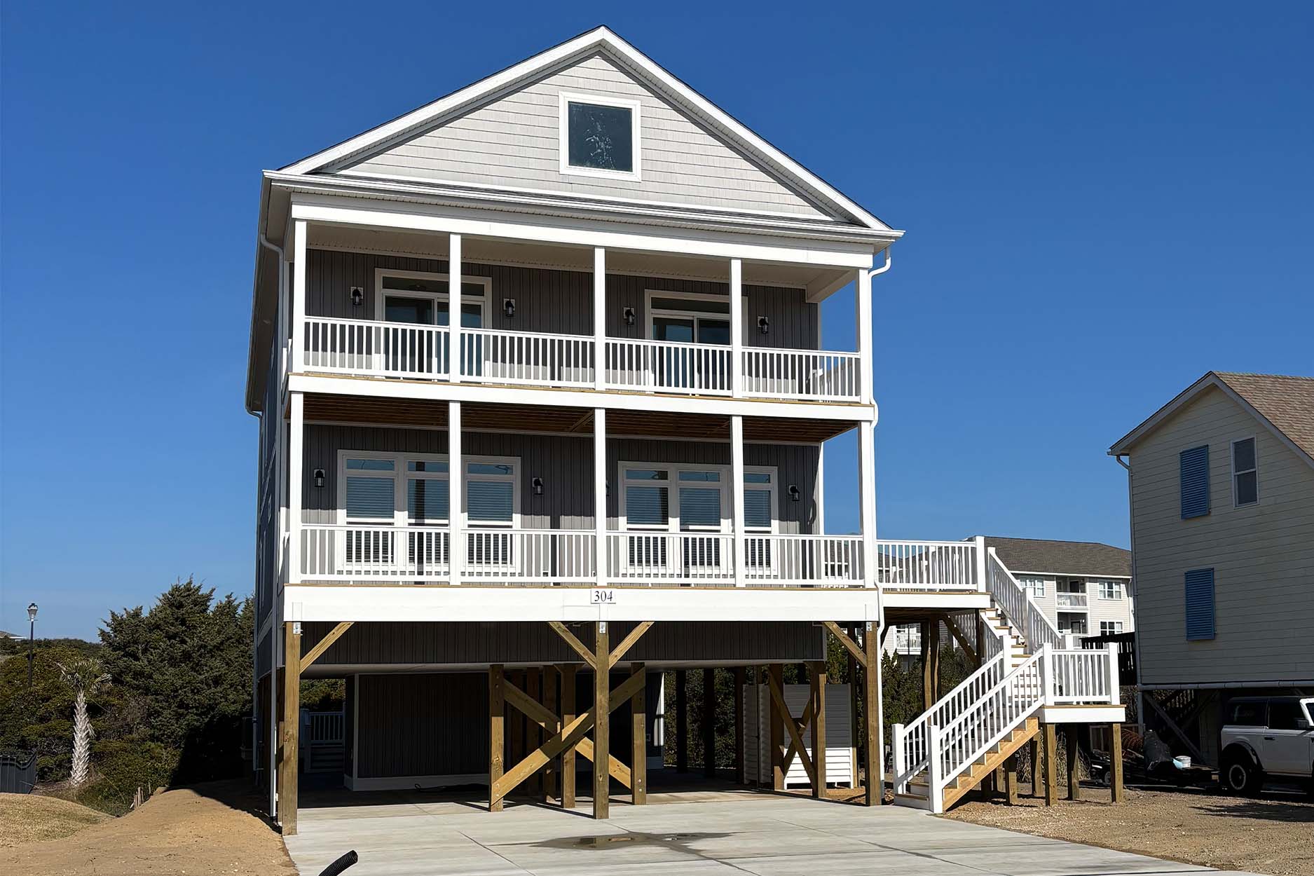 A three-story gray modular home on stilts with two levels of covered porches and an exterior staircase.